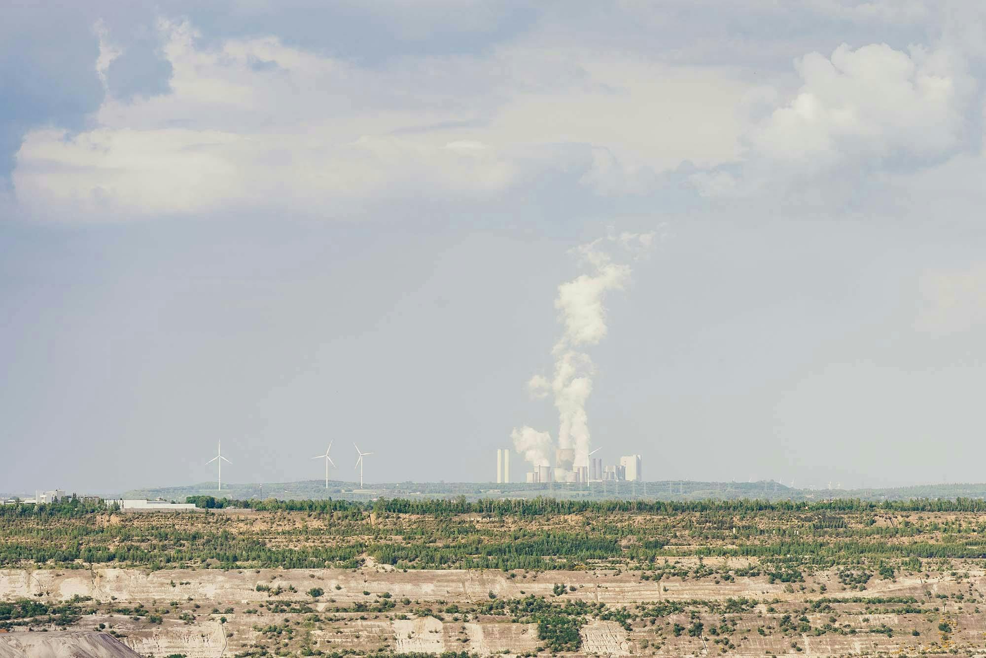 Rauch steigt aus Fabrik-Schornsteigen aus. Davor sind Windräder und eine trockene Landschaft zu sehen. (@barnimages)